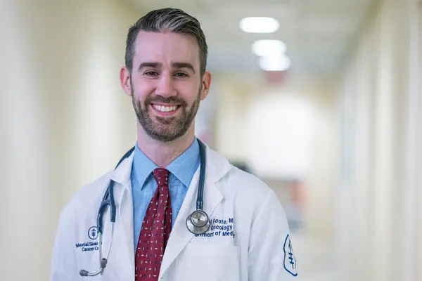 Dr. Michael Foote, with a stethoscope and wearing a doctors’ white coat, is smiling and standing in an MSK hallway.  