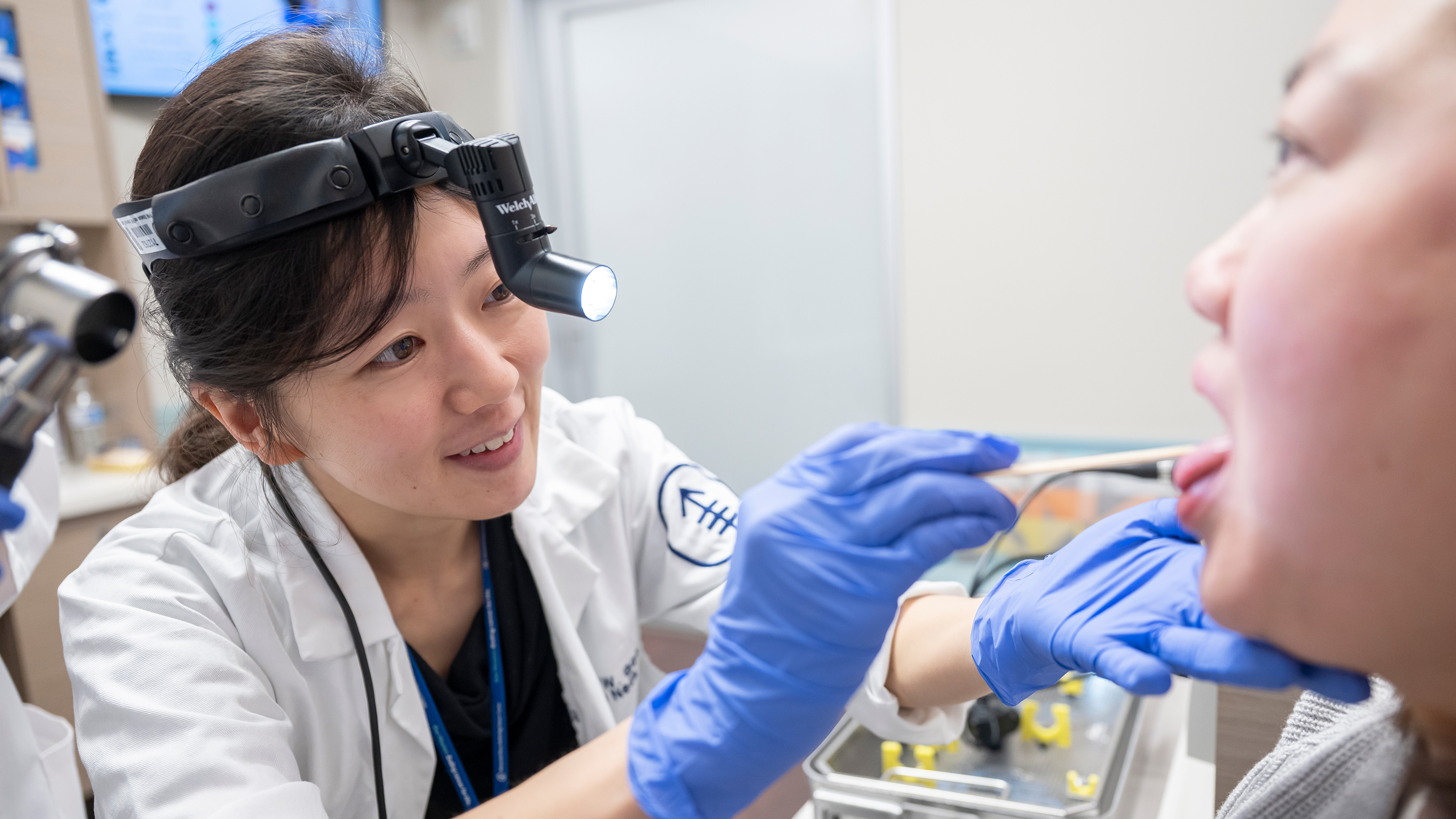 Head and neck surgeon Dr. Rebecca Gao, wearing a headlamp, uses her gloved hands and a tongue depressor to examine her patient’s mouth.  