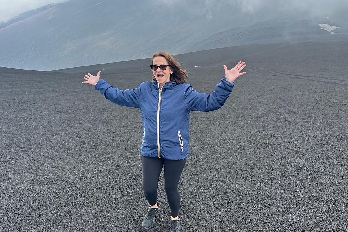 Woman standing on mountain in blue windbreaker with arms spread in celebration. 