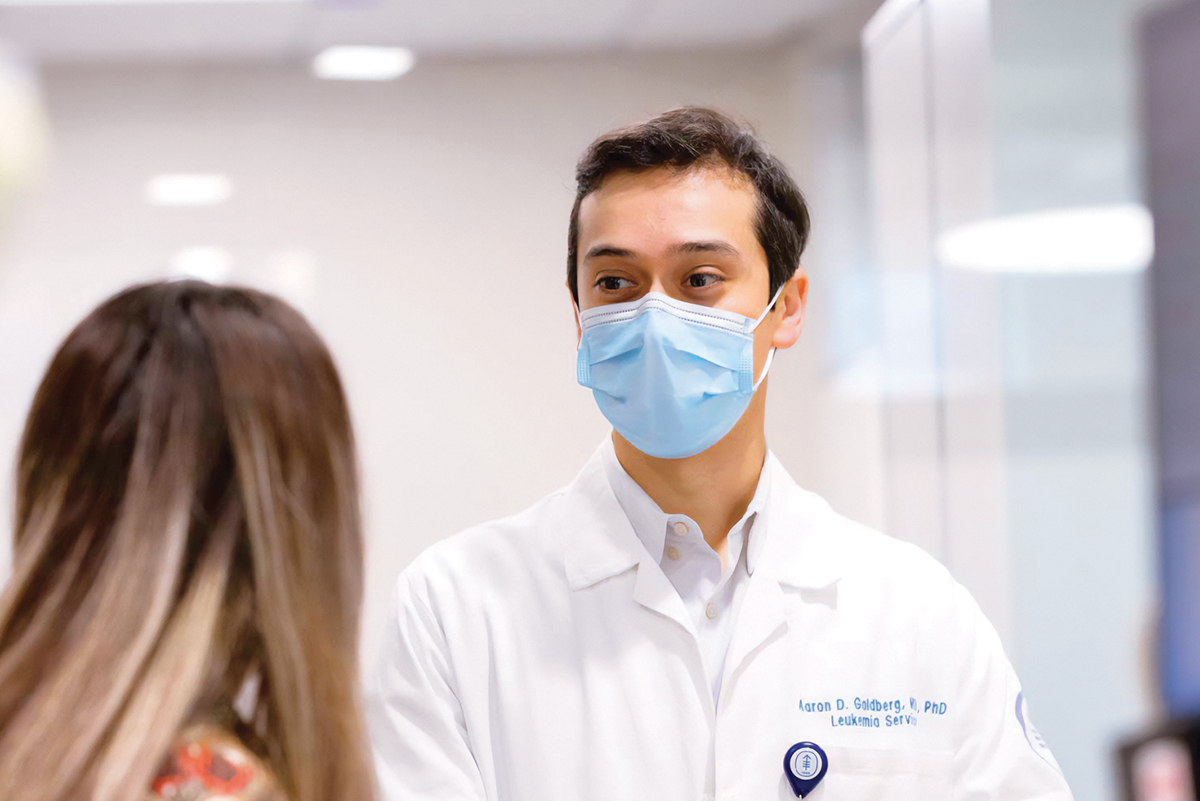 Photo of a man wearing a white coat and surgical mask, speaking to a woman whose face is not seen.