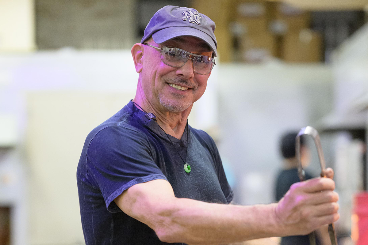 Man smiling at camera, holding a glassblowing tool.