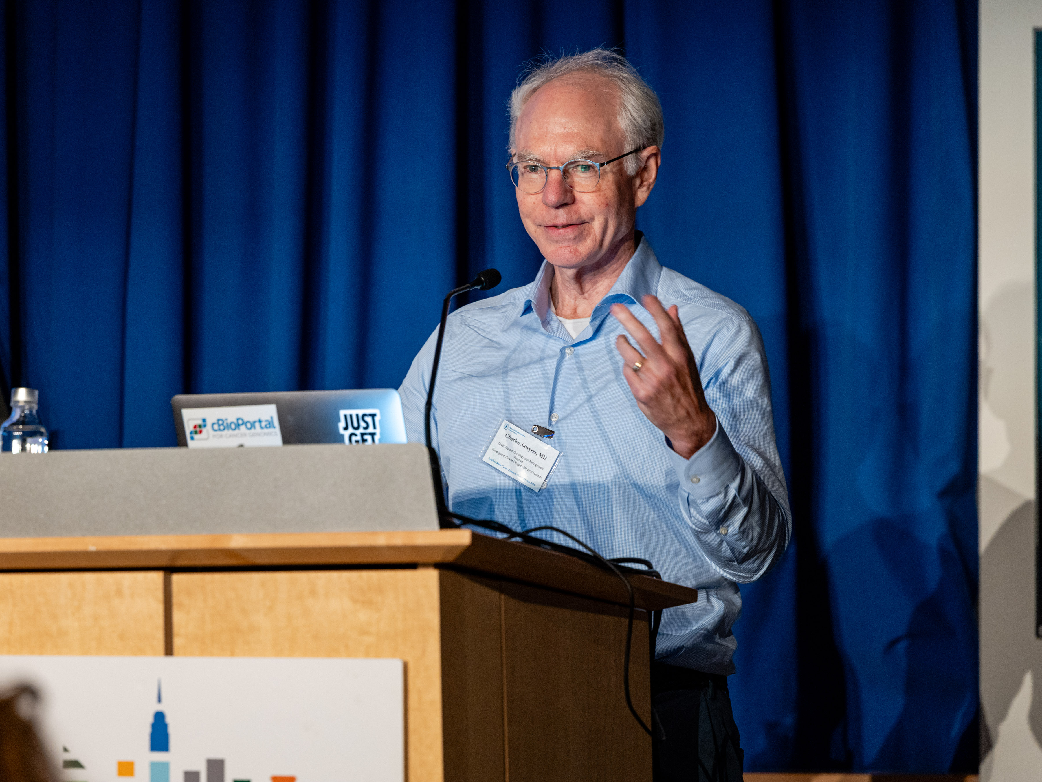 Dr. Charles Sawyers stands at a podium while giving his talk, one of his hands are raised and he's smiling.