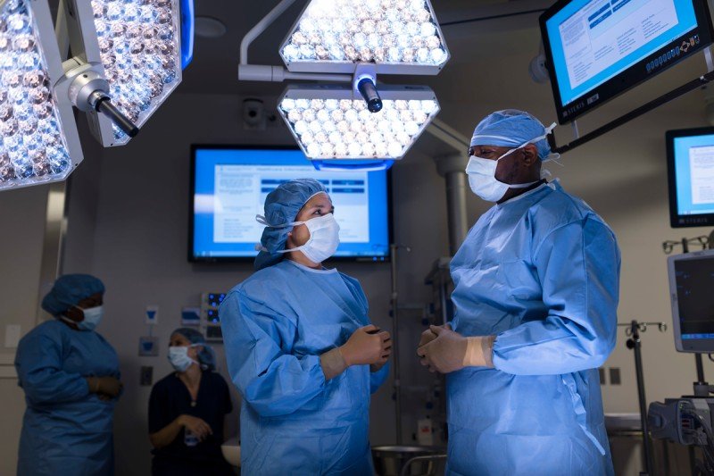 Two people in surgical scrubs stand in an operating room underneath large light panels and screens with medical information.