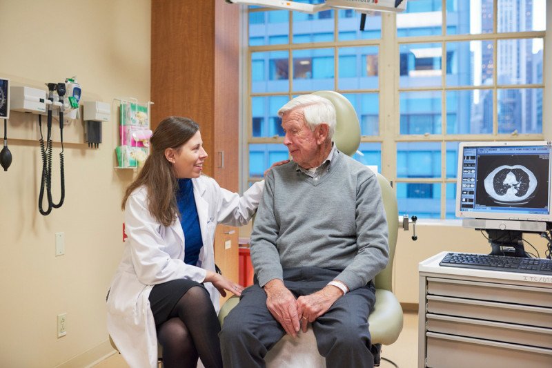 A young female doctor in a white coat sits with an older male patient in an exam room