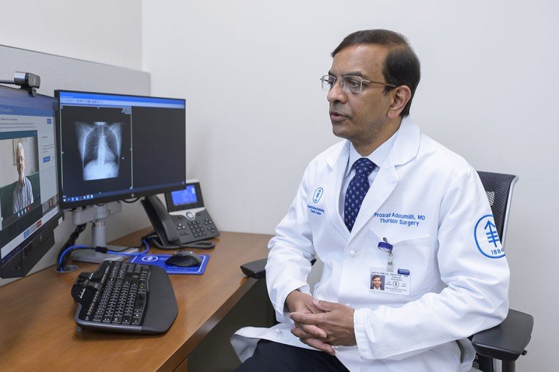 El Dr. Prasad Adusumilli sentado frente a la computadora. En el monitor, se puede ver la cara y una imagen del torso de un paciente.