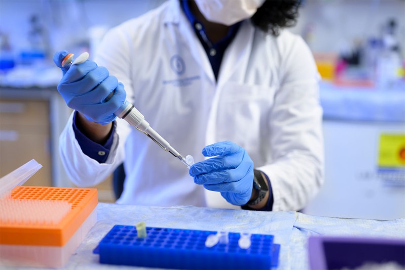 Person in white lab coat using a pipette with a tiny test tube.