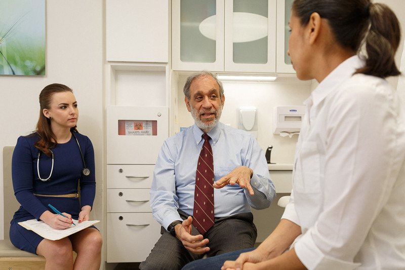 Doctor seated in center speaks with patient (on exam table on right) while assistant seated on left observes.