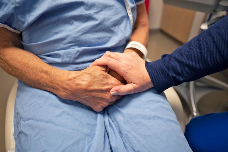A doctor holds the hand of a patient, who is dressed in a hospital gown