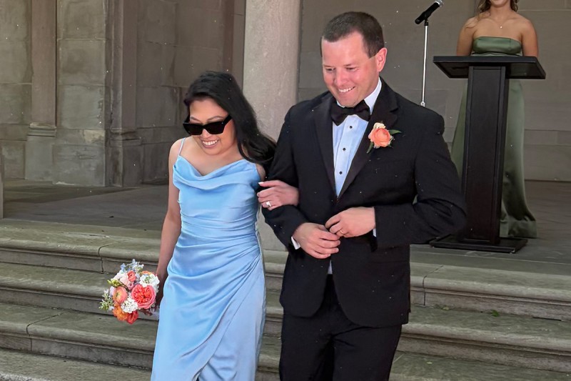 Christina and Kevin walk down stairs. Christina is wearing a blue dress and carrying a bouquet. Kevin is wearing a tuxedo.