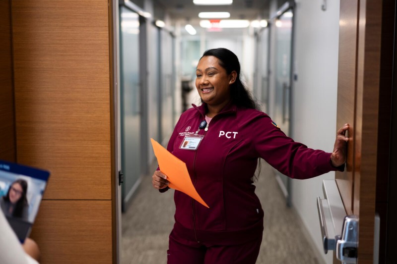 Una técnica de atención al paciente con un uniforme rojo oscuro que abre una puerta y saluda al paciente en un pasillo bien iluminado.