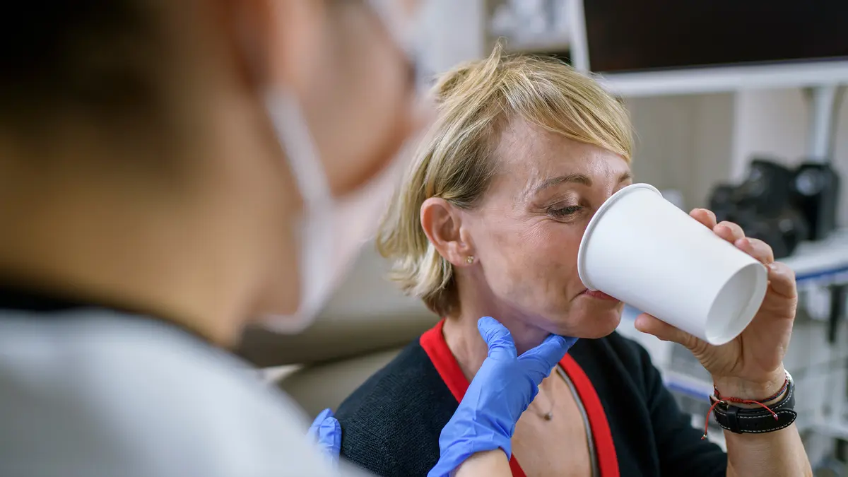 During an exam, an MSK speech pathologist holds a gloved hand to the throat of a patient who drinks from a plastic cup. 