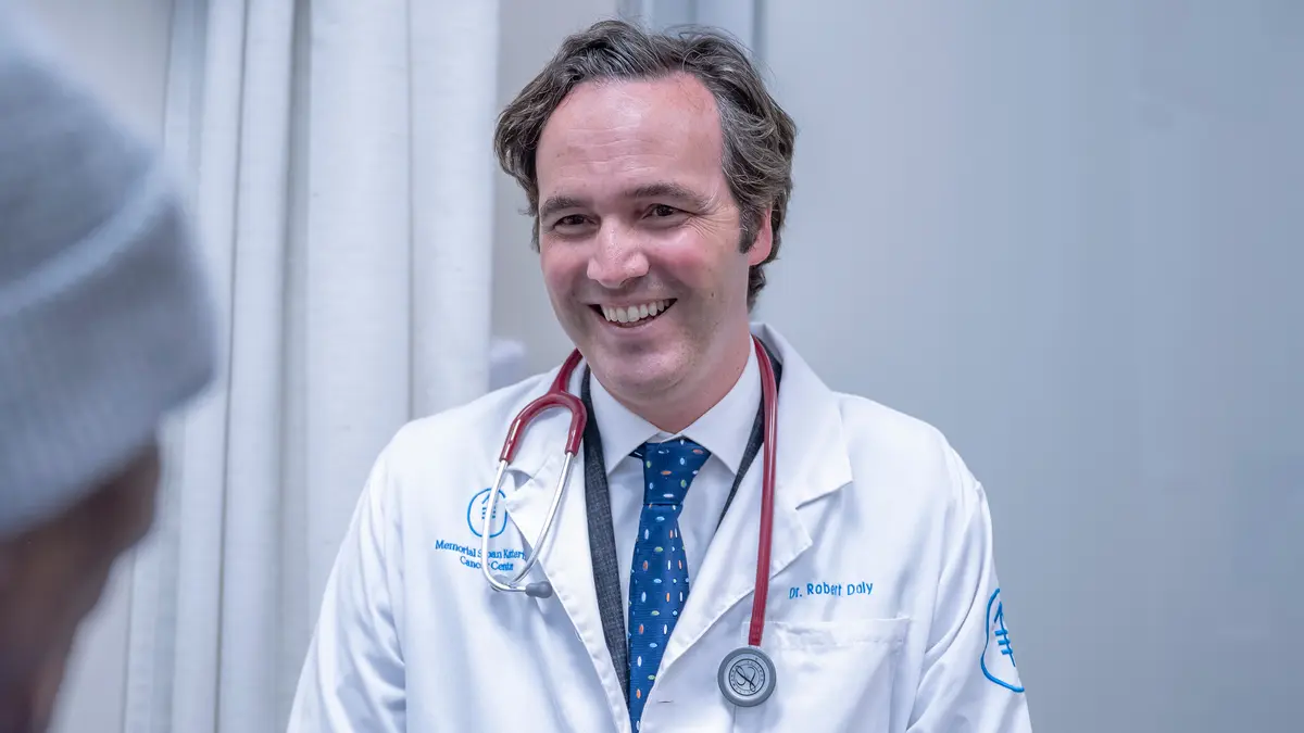 MSK medical oncologist Dr. Robert Daly is seen smiling with a stethoscope draped around his neck while he listens to a patient.