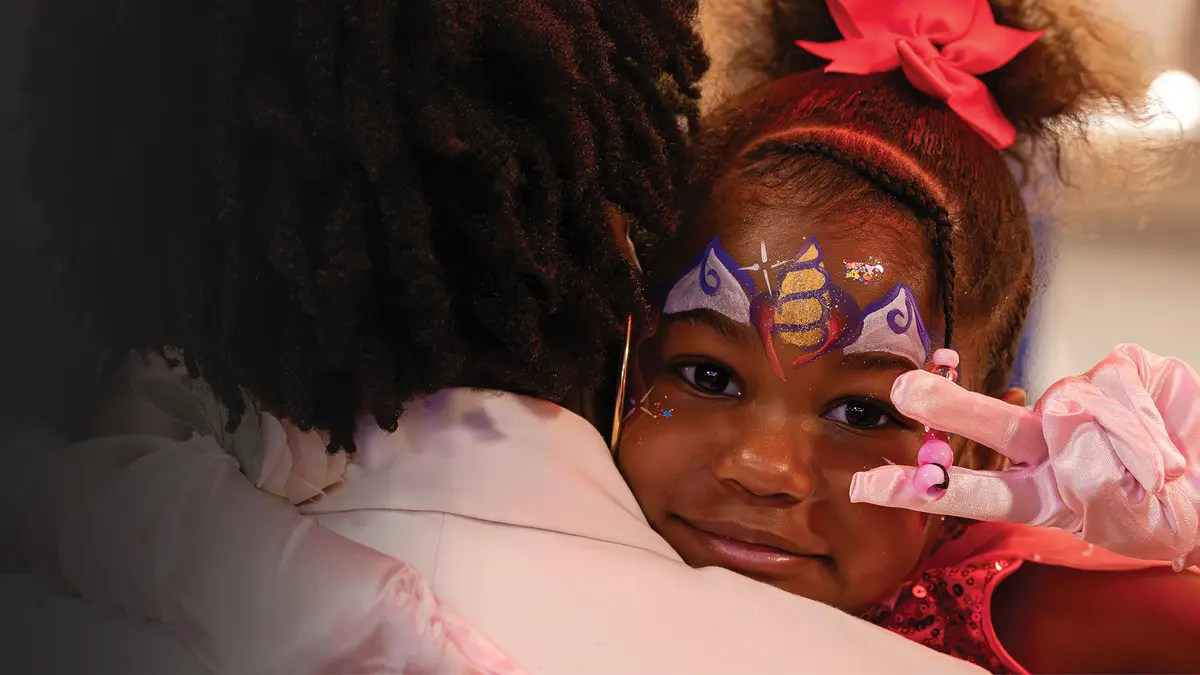 A young girl wearing colorful face paint for the MSK Kids prom looks over her mother’s shoulder making a peace sign and smiling.