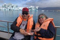 A man and a woman sit on a boat wearing life jackets and holding a large icicle. An iceberg sticking out of the water is behind them.
