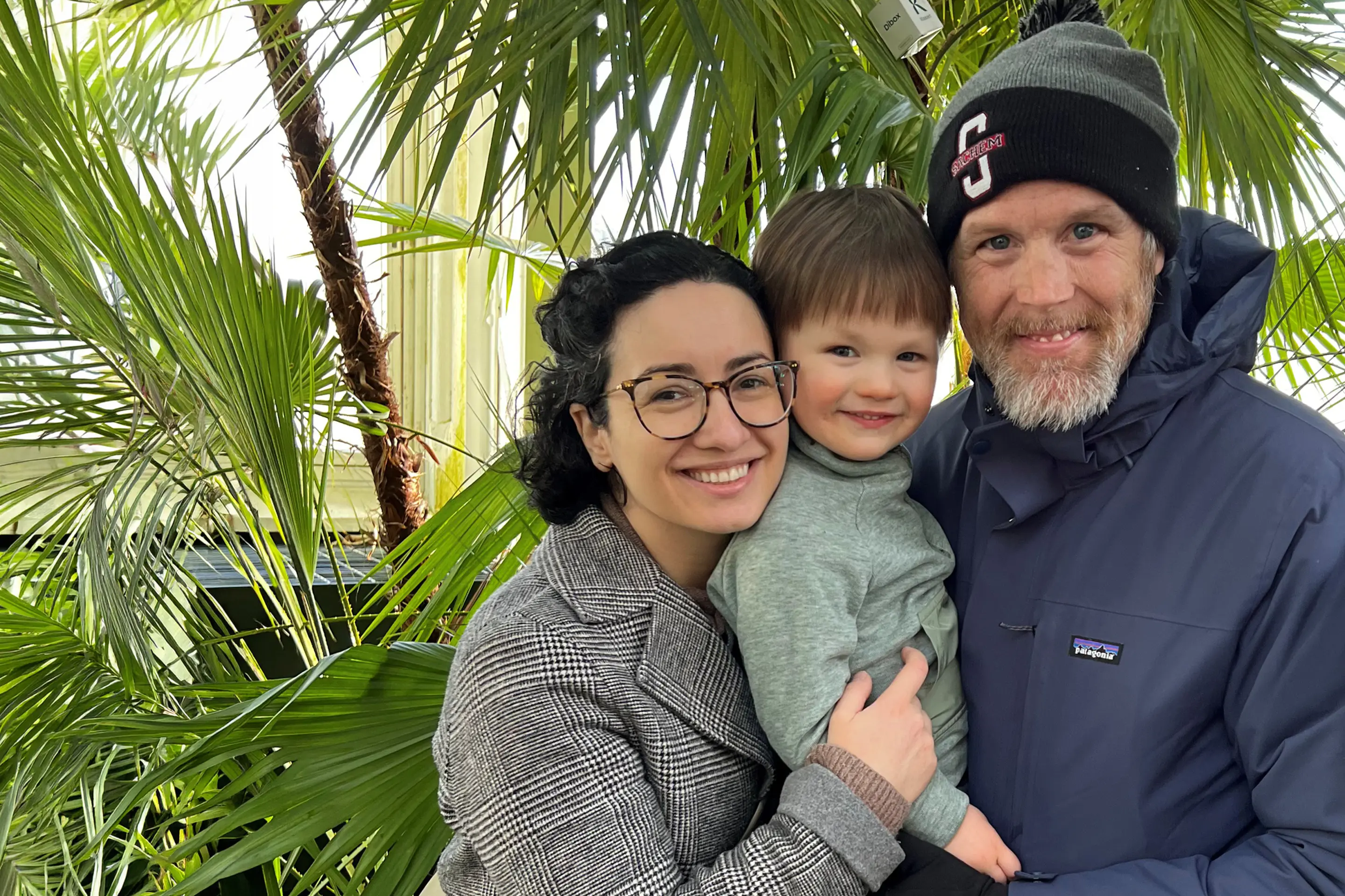 Kevin Tougher and his wife, Maria, holding their son, Theo, surrounded by trees outdoors. 