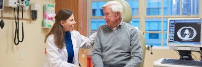 A young female doctor in a white coat sits with an older male patient in an exam room