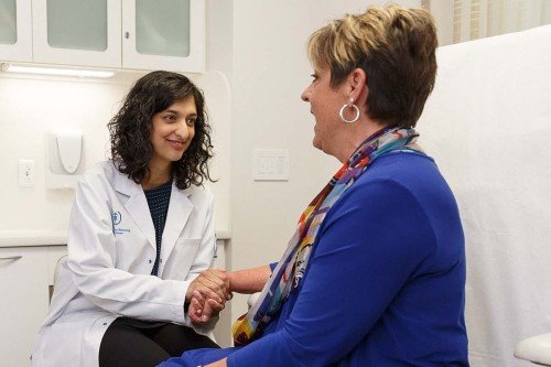 Female doctor in lab coat on left speaks with patient wearing blue top.