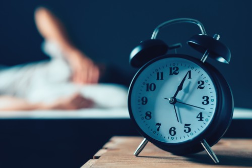 Alarm clock in foreground with person (out of focus) in bed in the background.